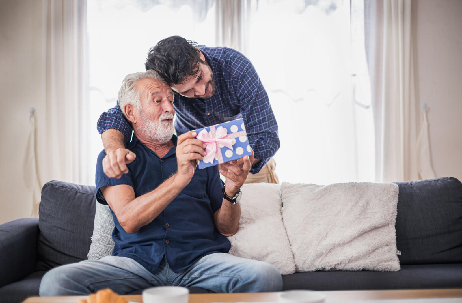 An older parent with dementia looking at a gift with curiosity while their adult child hugs them from behind and laughs.