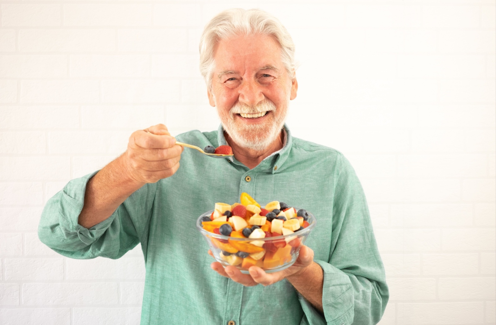 A smiling older adult holding a bowl of cut fruit in his left hand and a spoonful of fruit in the right hand.