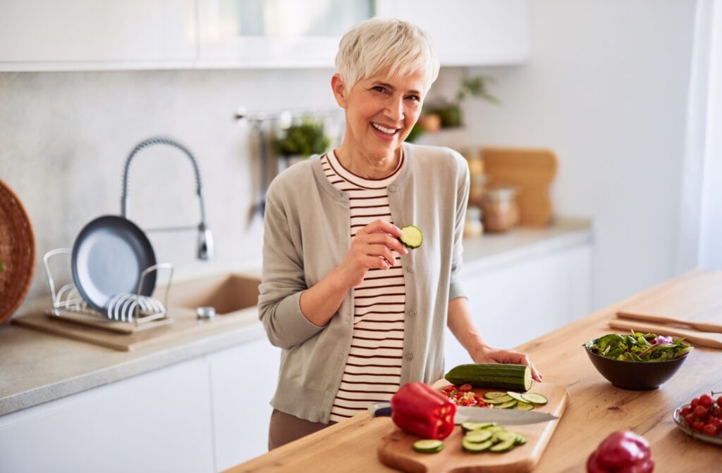 A smiling older adult holding a slice of cucumber in her right hand over a kitchen counter with fresh vegetables.