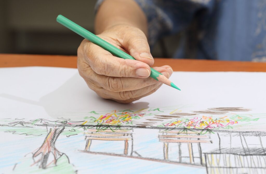 The elderly individual's hand, delicately wielded a green-colored pencil, sketching an outdoor landscape on a drawing pad.