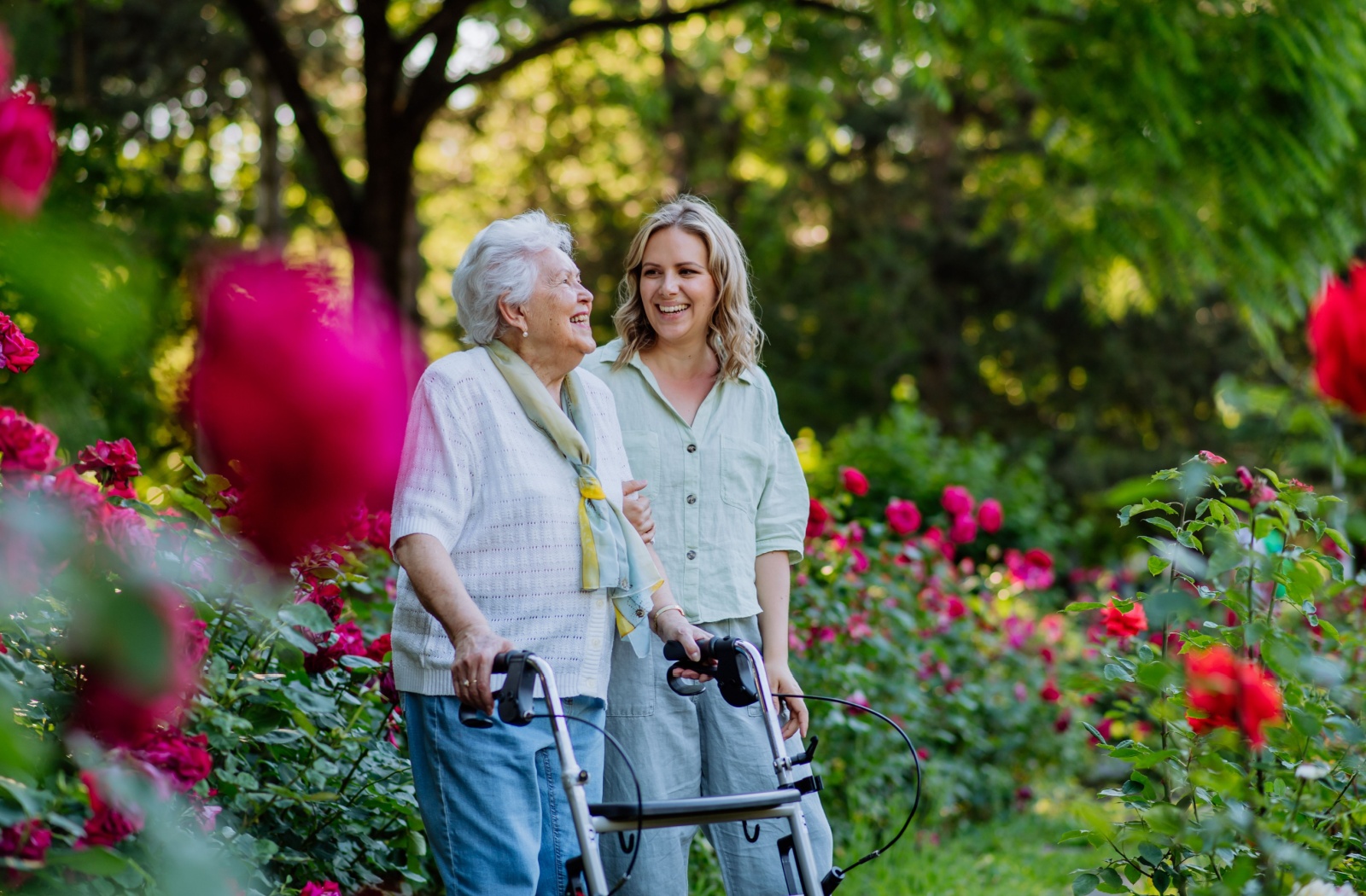 An older woman with a walker walking through a rose garden with her adult daughter and laughing.