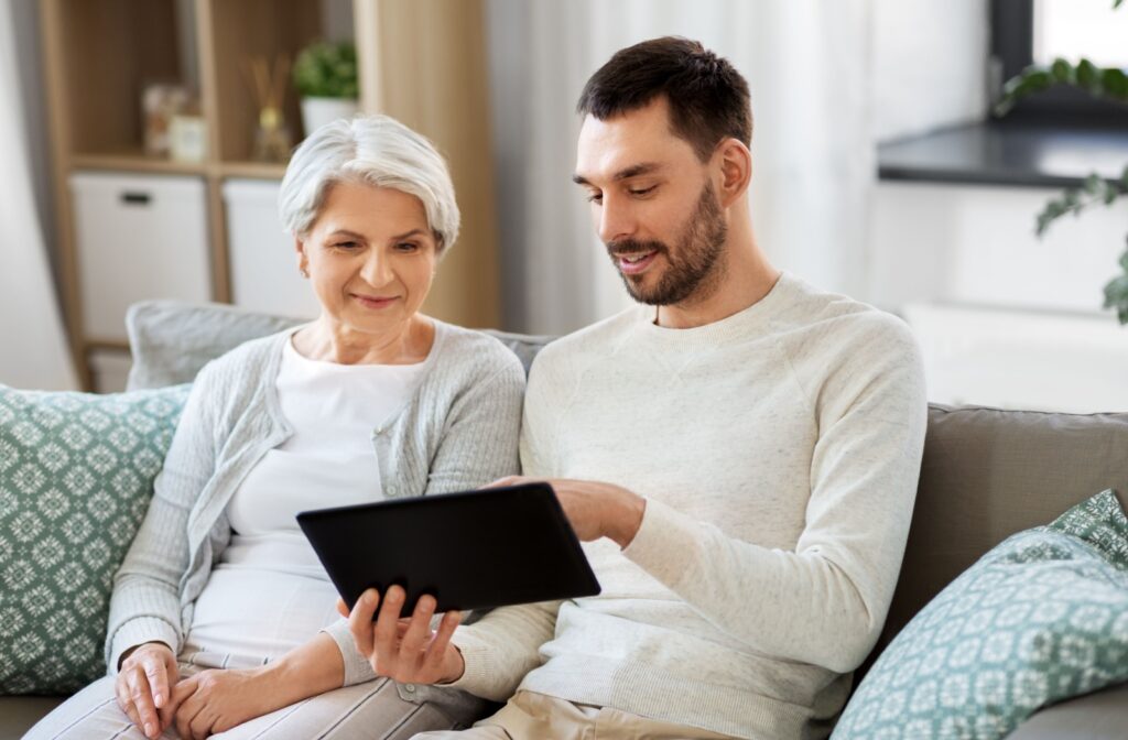 A young man sitting on a couch with his older mother and showing her something on a tablet.